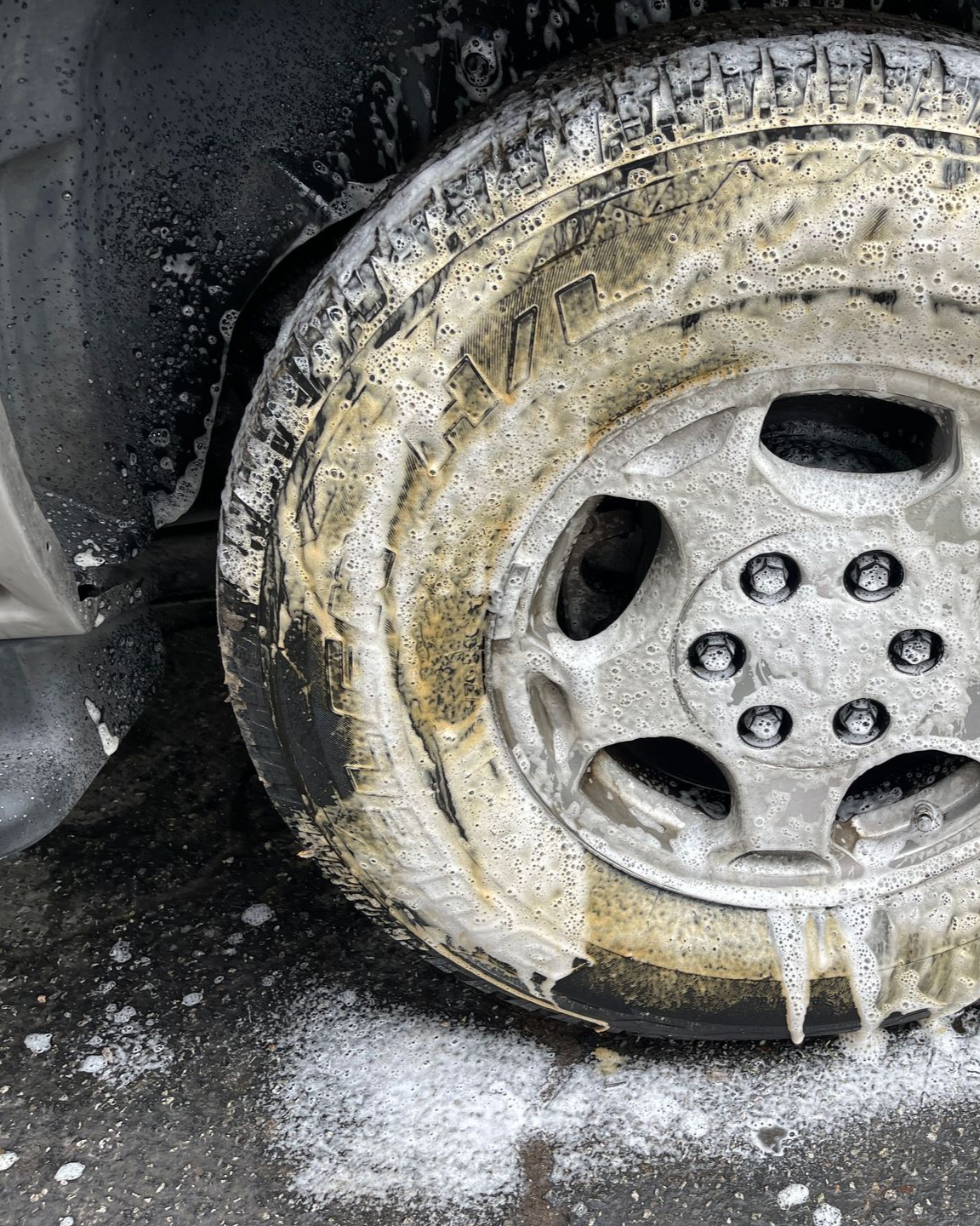 Close-up of a dirty car wheel with white foam and grime washing off the tire and rim.