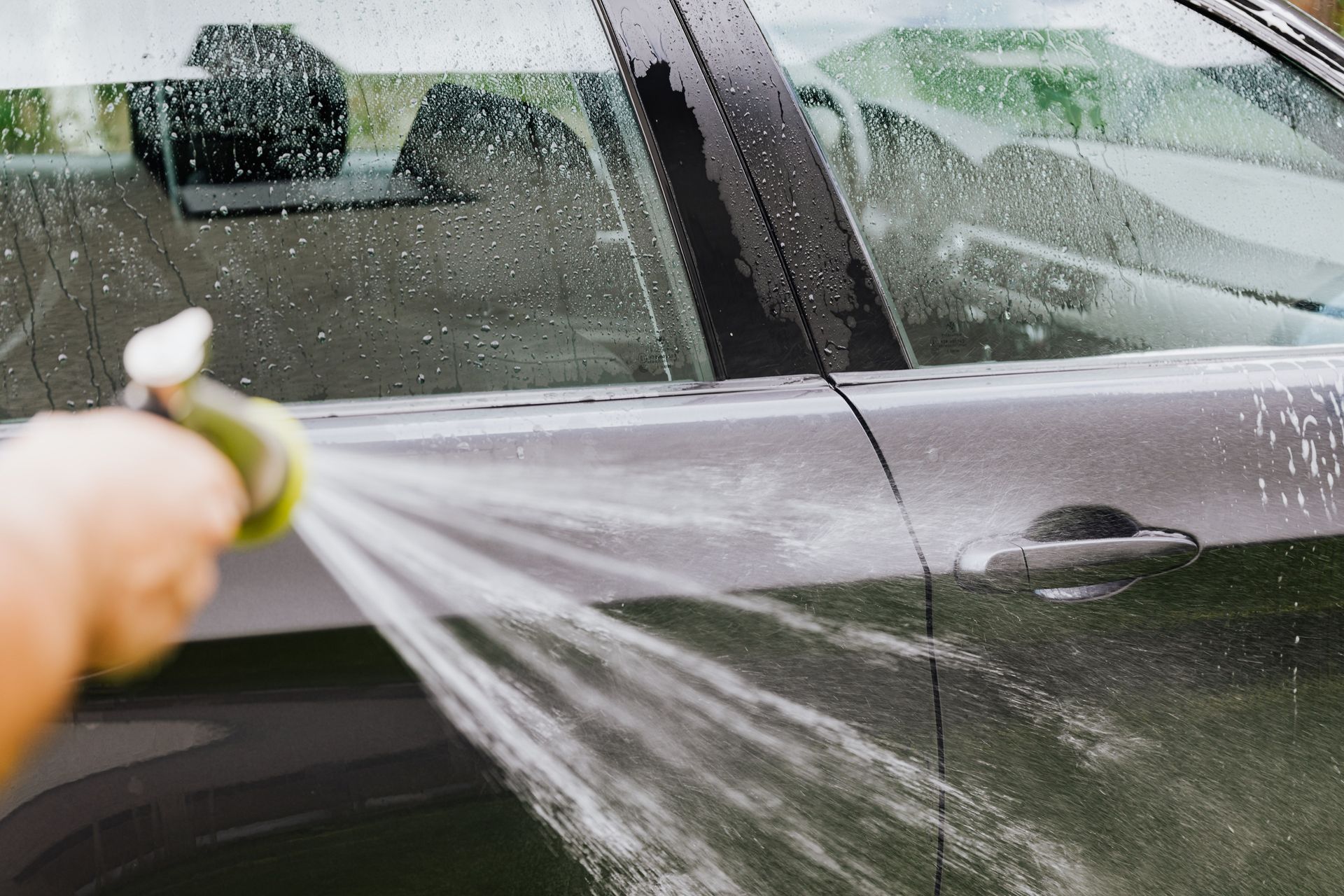 A hand sprays water on a black car door to rinse it clean.