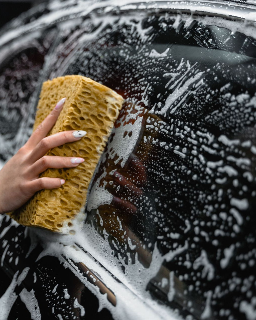 Hand washing a black car with a yellow sponge and white soap suds