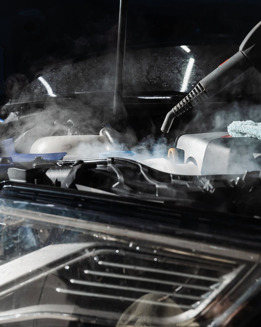 Steam rises from a car engine bay while a person in a black glove inspects it with a tool.