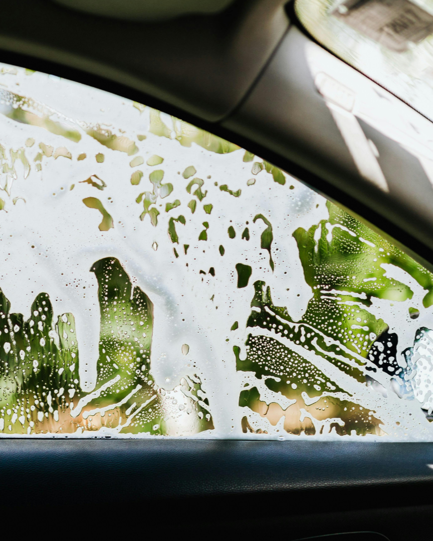Car window covered in soap suds and water droplets, with green foliage visible outside
