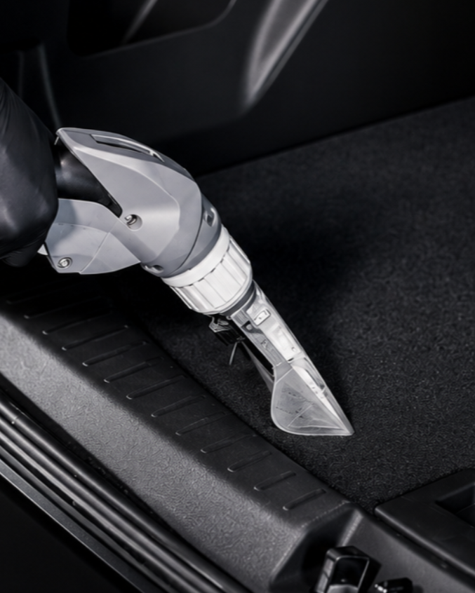 A robotic vacuum cleaner cleaning black carpet in a car trunk.