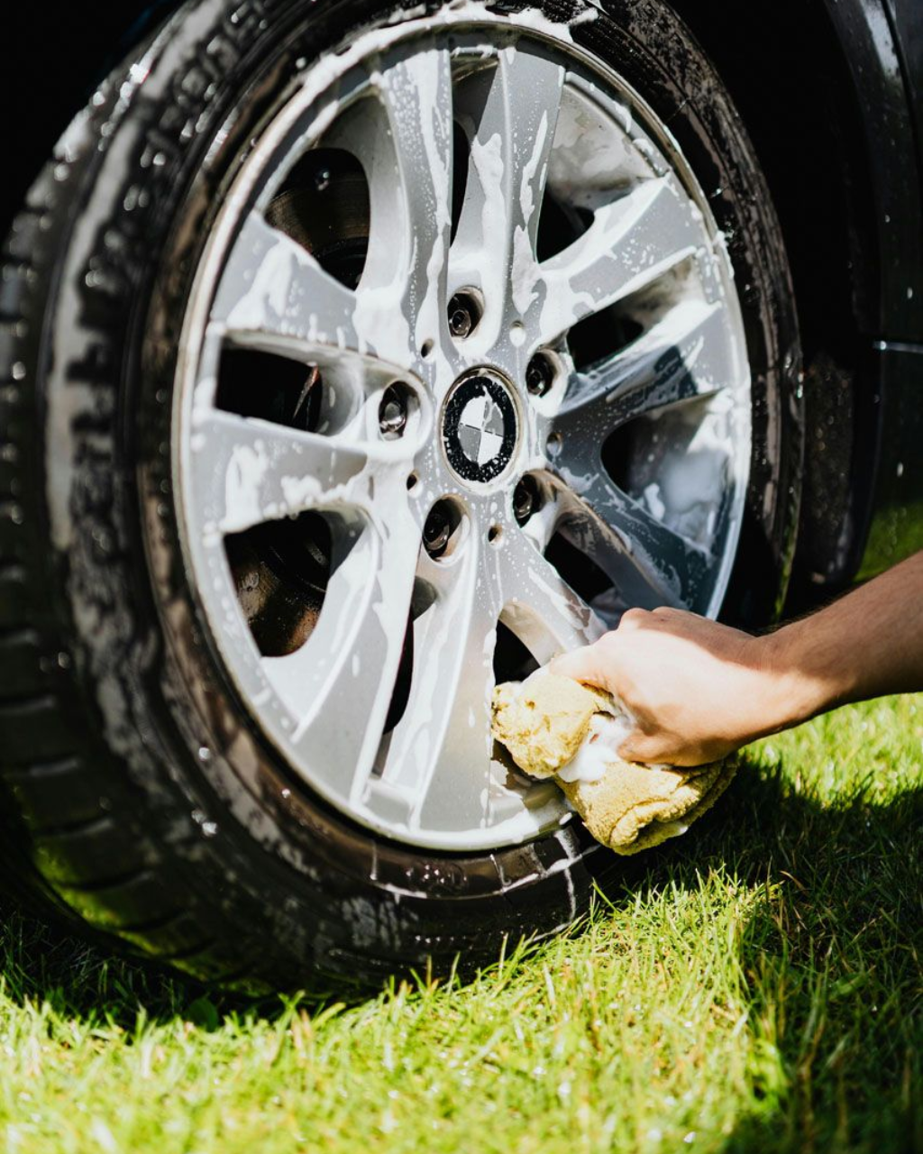 Hand washing a dirty car wheel with a sponge on grass.