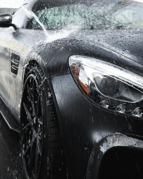 Close-up of a black sports car being washed, with water spraying over the hood and windshield.