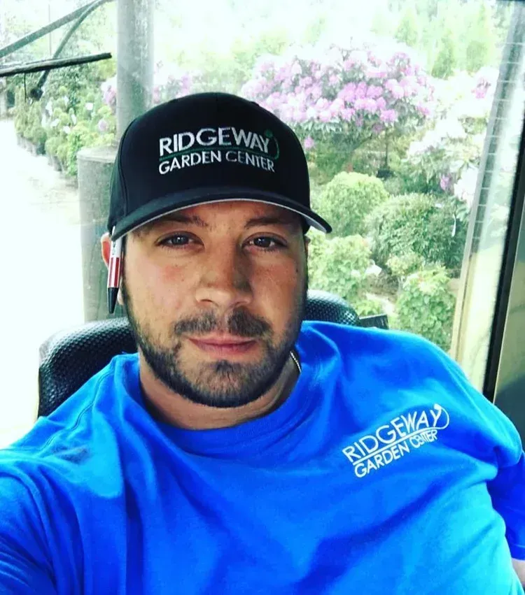 Man wearing a Ridgeway Garden Centre cap and shirt, seated indoors with a garden view behind him