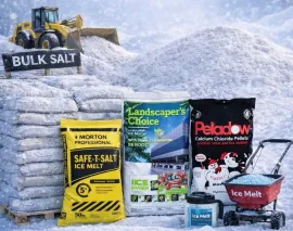 Bulk salt and ice melt products stacked in a snowy lot with a snowplow and wheelbarrow.