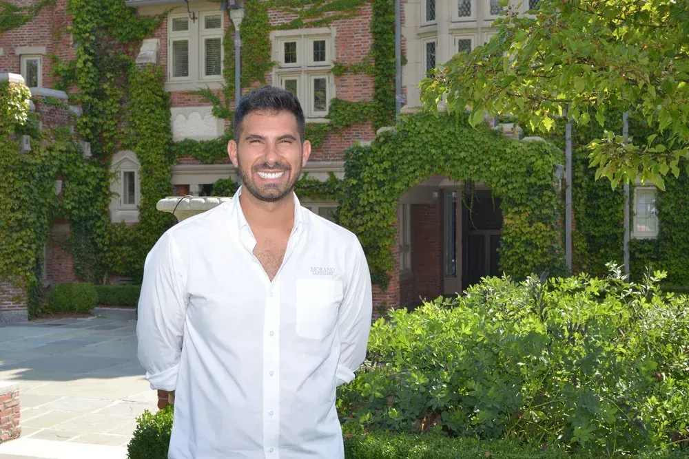 Smiling man in a white shirt standing outdoors in front of ivy-covered buildings and greenery