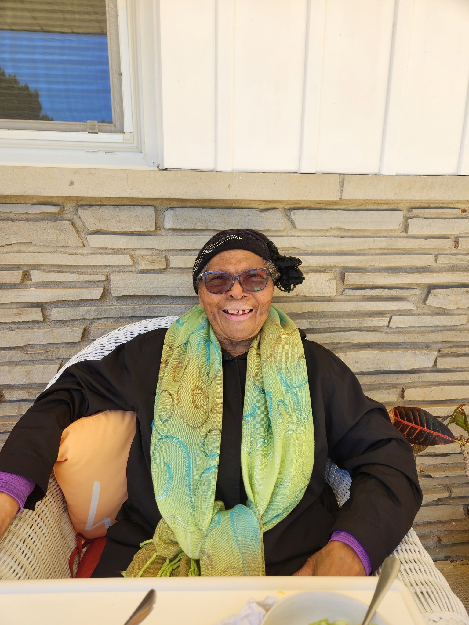 Person seated on a porch, wearing a black outfit, purple gloves, and a green patterned scarf, smiling.