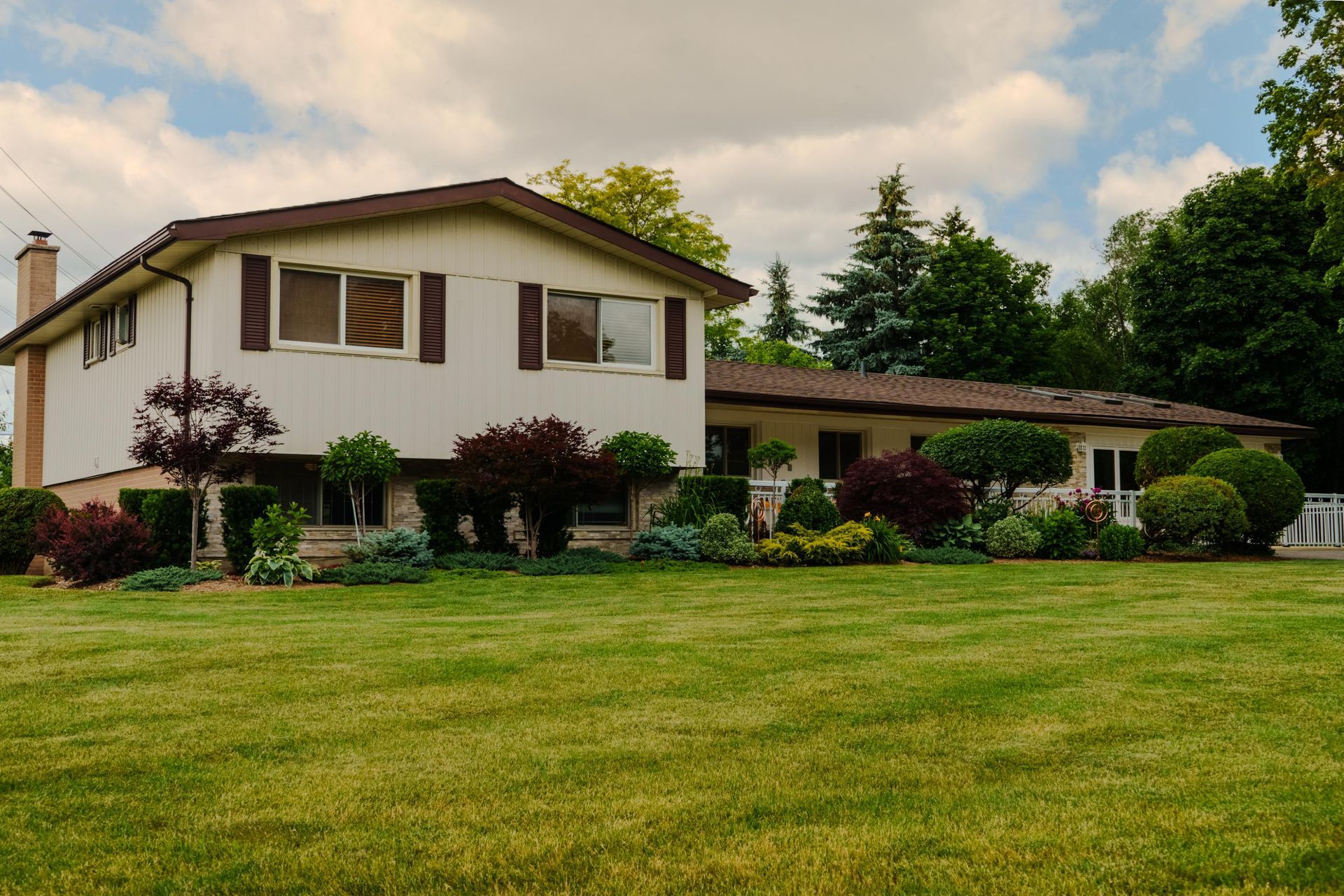 Suburban house with white siding, brown roof, and a large green lawn under a partly cloudy sky