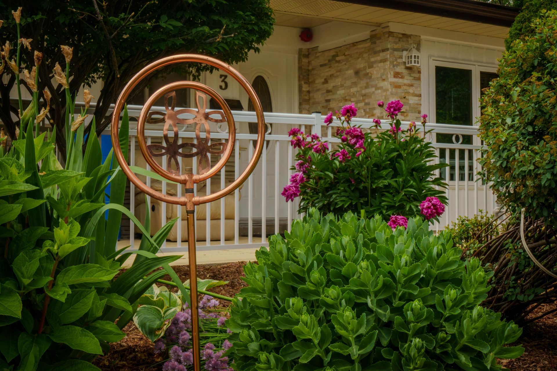 Copper wind sculpture among lush green plants and pink flowers in a garden yard