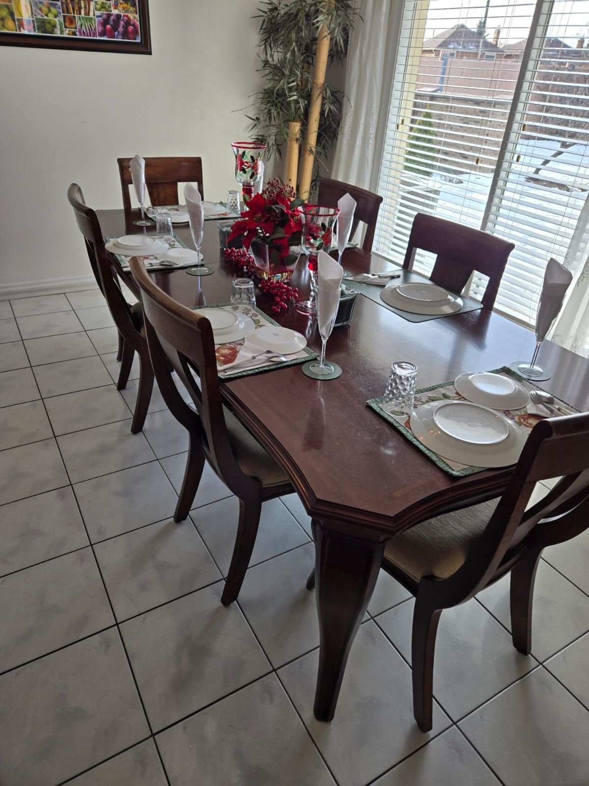 Set dining table with white plates, glasses, and red floral centerpiece beside a window.