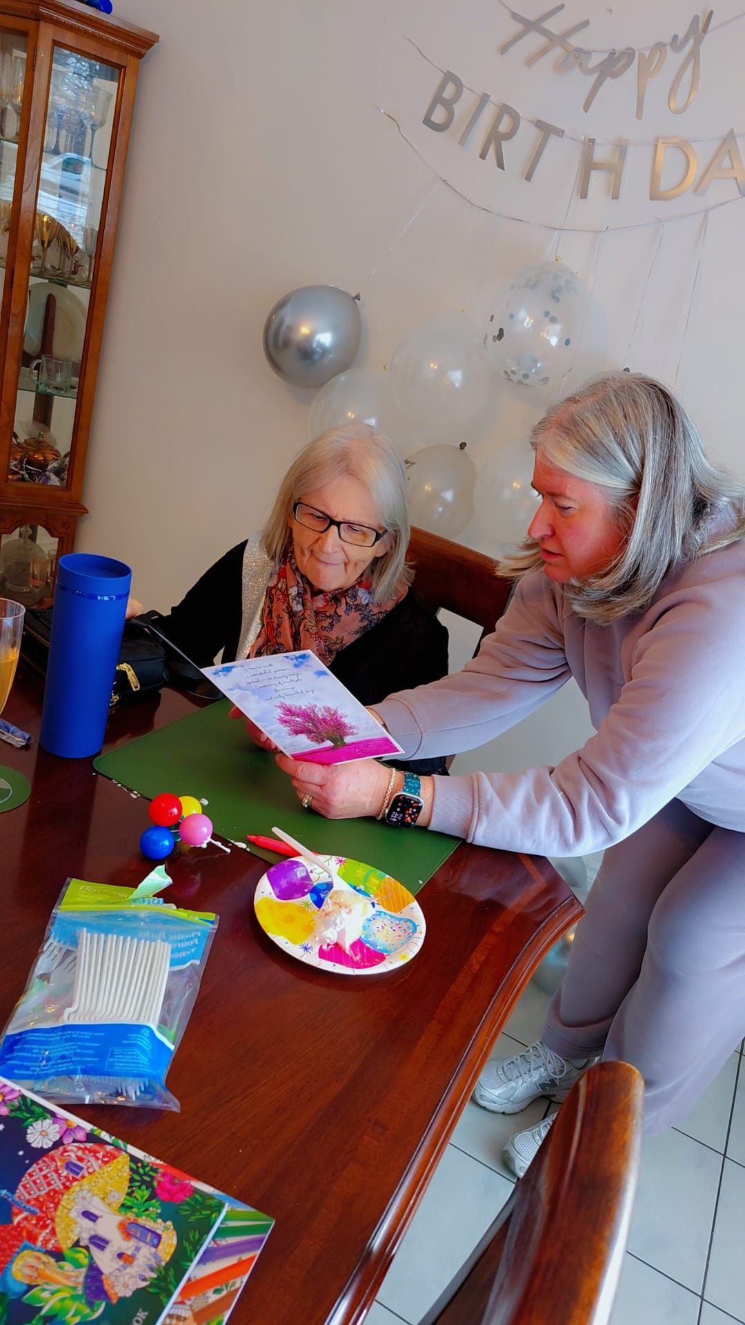 Woman helping child open a birthday gift at a decorated table with balloons and party items