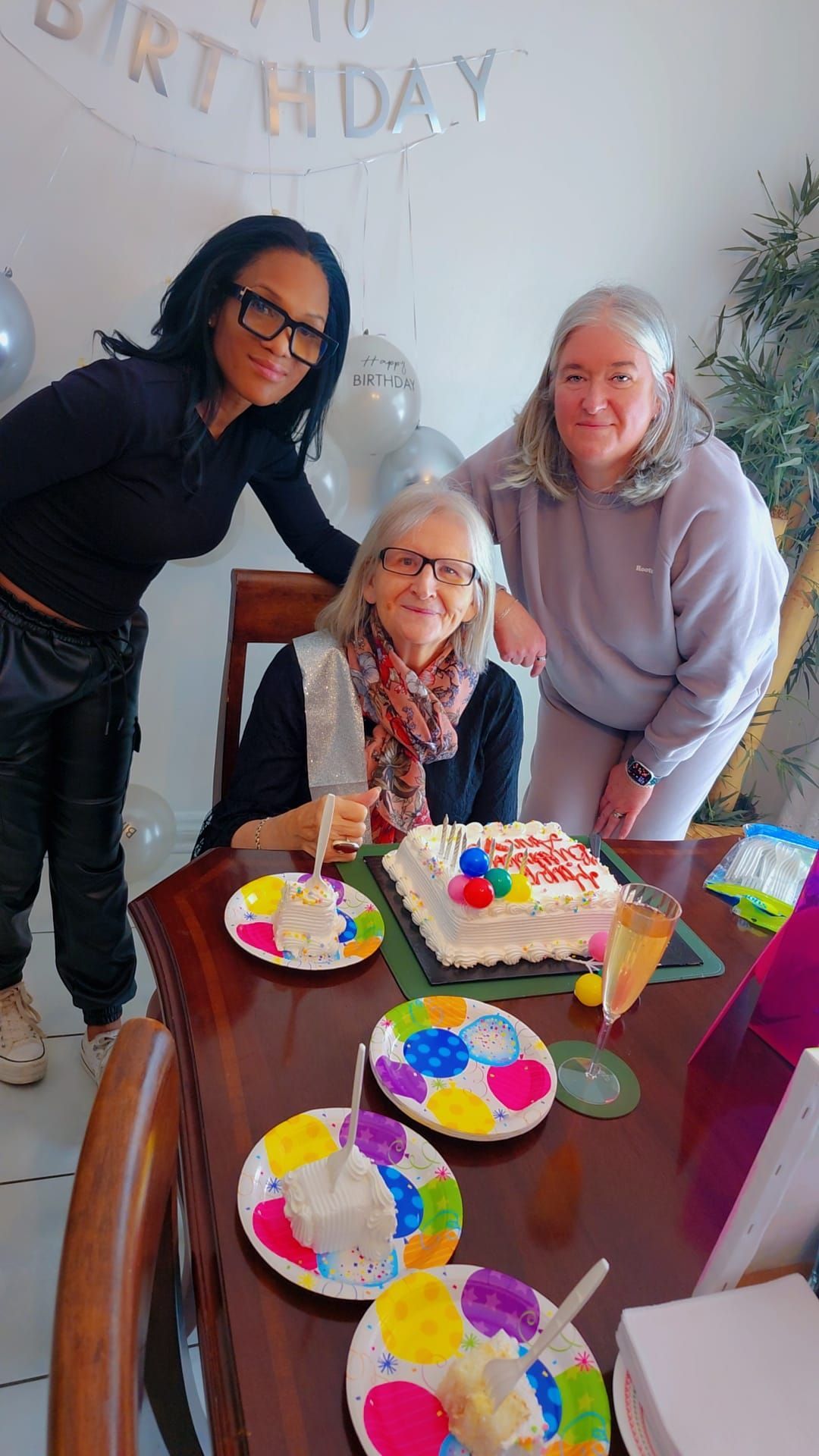 Three women pose at a birthday table with cake, colorful plates, and balloons.