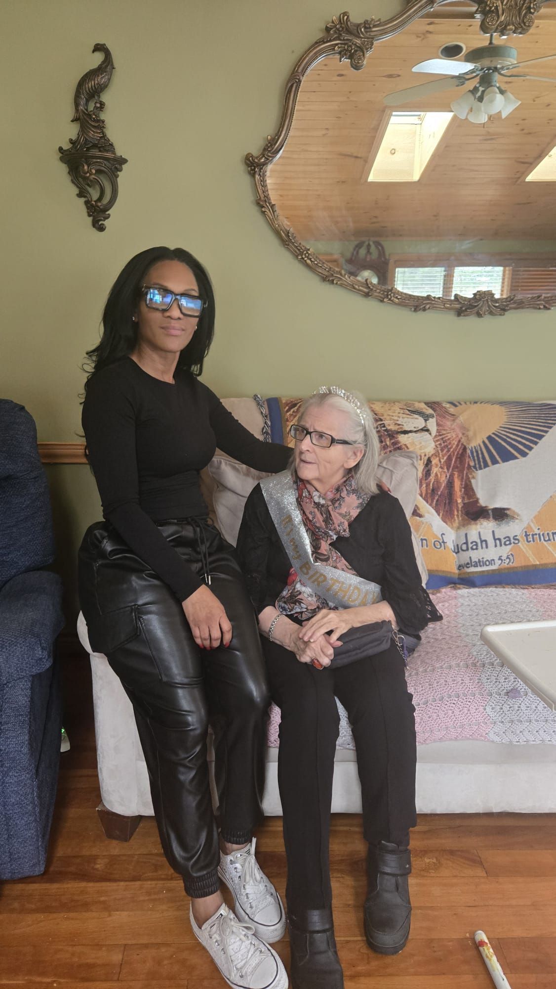 Two women sitting on a couch in a living room, one standing beside it, with a mirror on the wall.