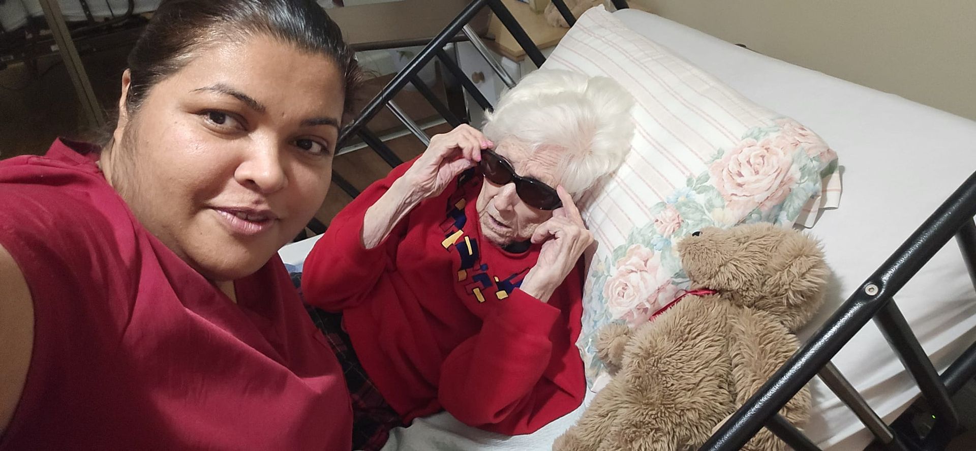 Woman in red top taking a selfie beside a child in red jacket and sunglasses on a bed