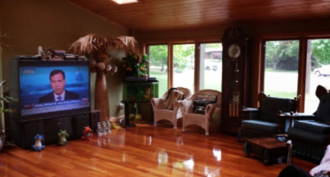 Living room with polished wood floors, TV, sofa, chairs, and large windows overlooking greenery