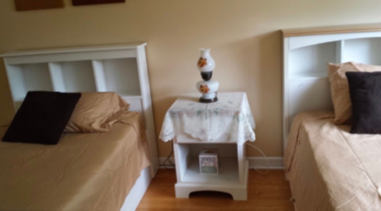 Two twin beds with beige bedding flank a small white nightstand in a bedroom.