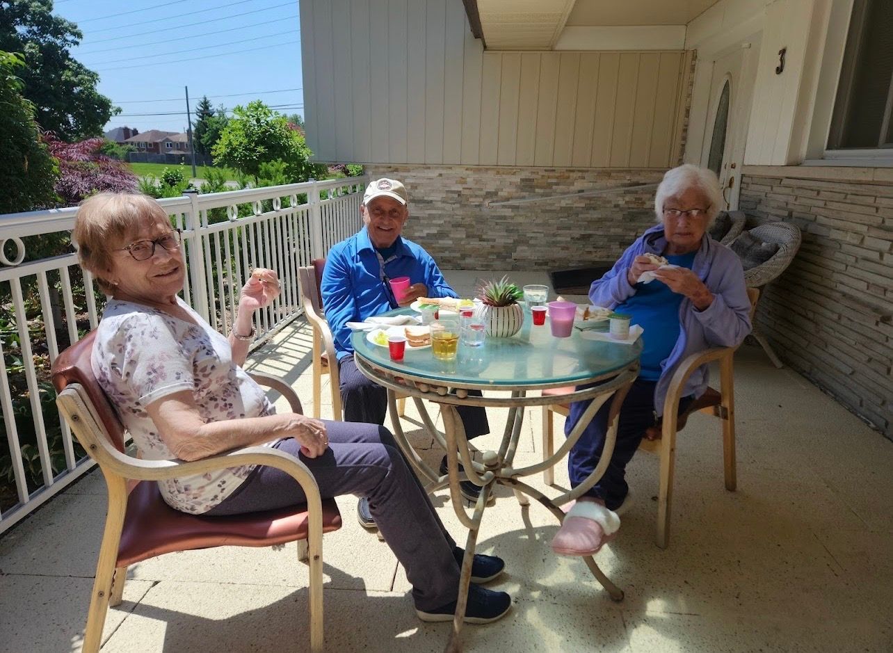 Three elderly people sit on a porch around a glass table, chatting in the sun.