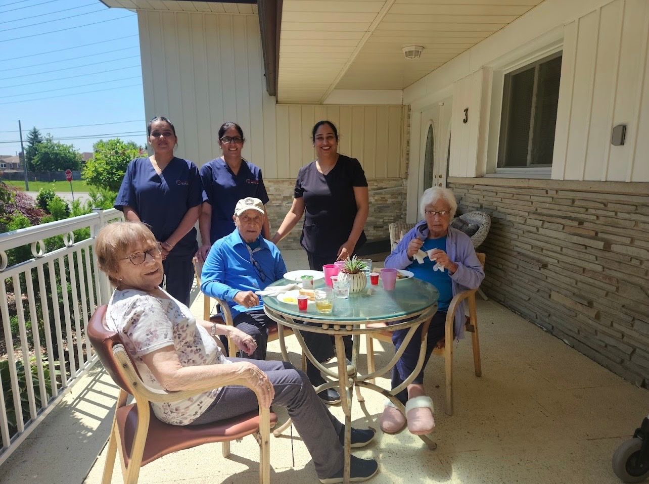 People seated around a patio table outside a house, with two standing nearby in bright sunlight