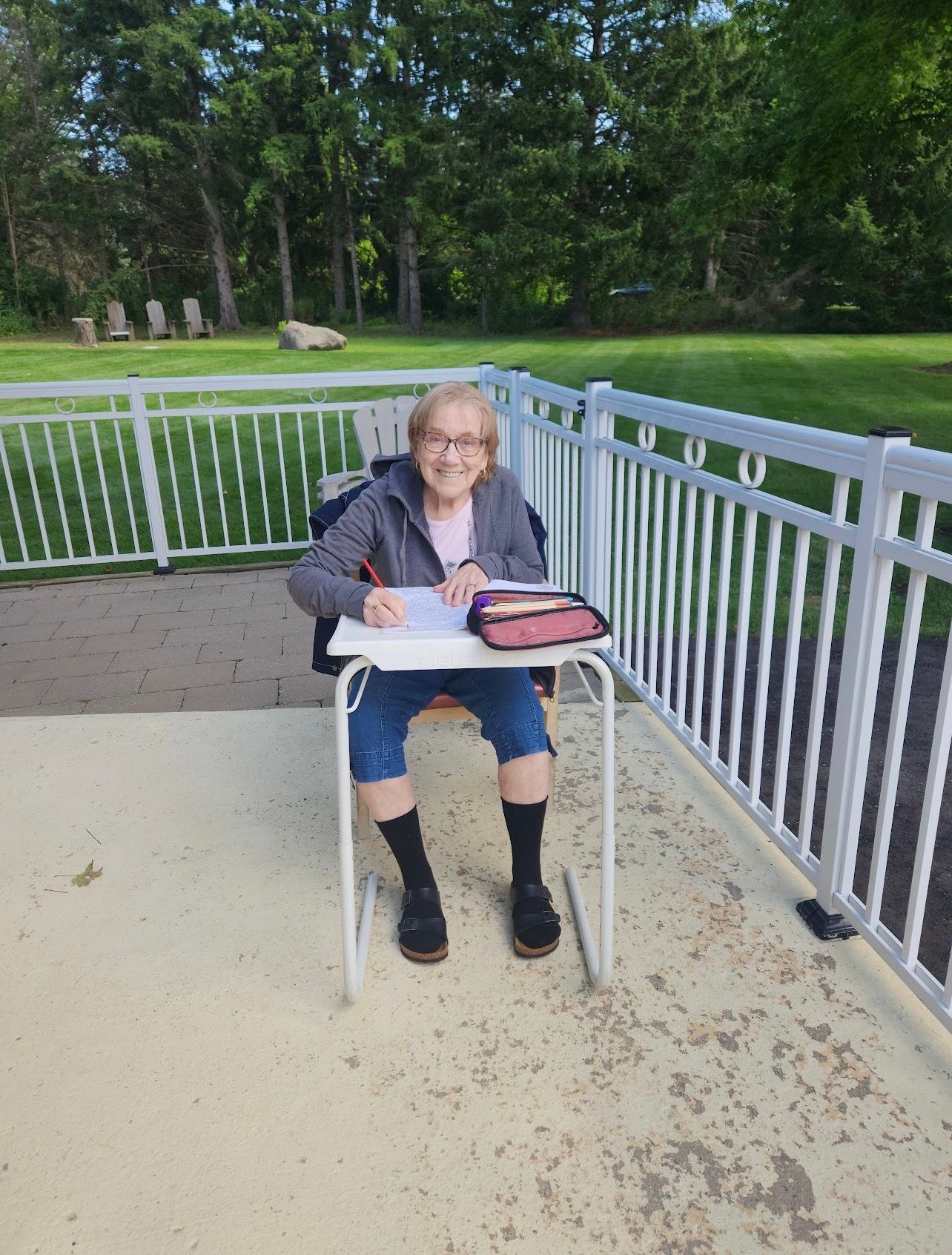 Person sitting at a small table on a porch, writing in a notebook beside a white railing and green lawn.