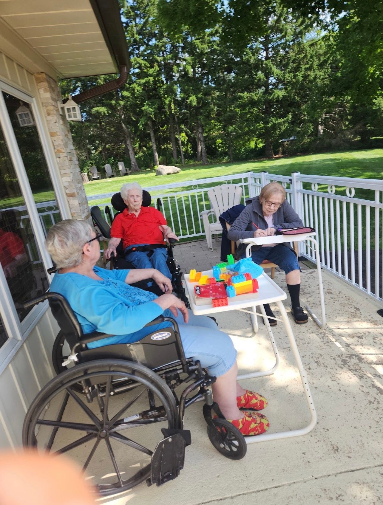 Three people sit on a porch with a table of colorful items, overlooking a green lawn.