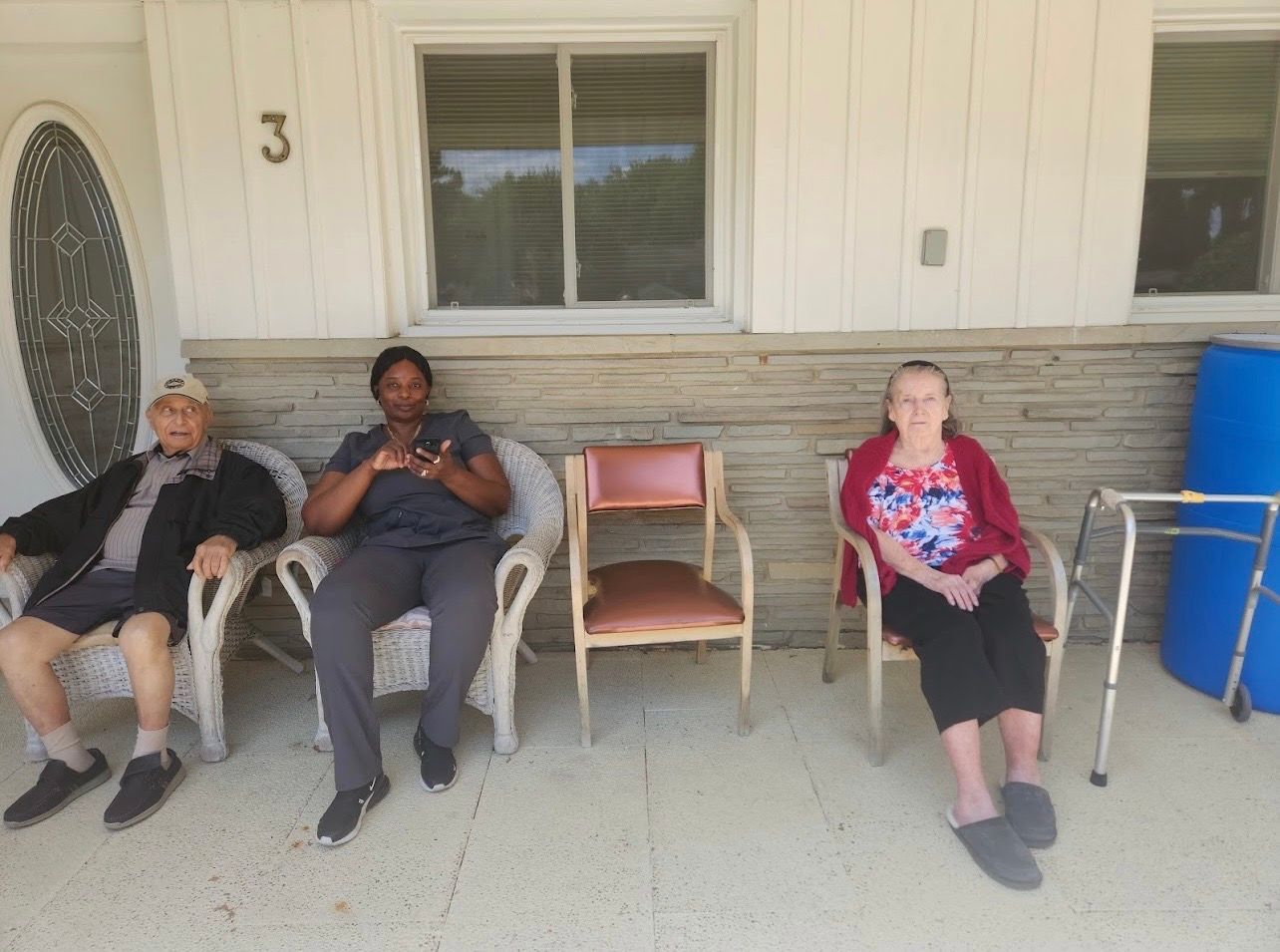 Three people sitting in chairs on a porch beside a house, with a red chair and blue bin nearby