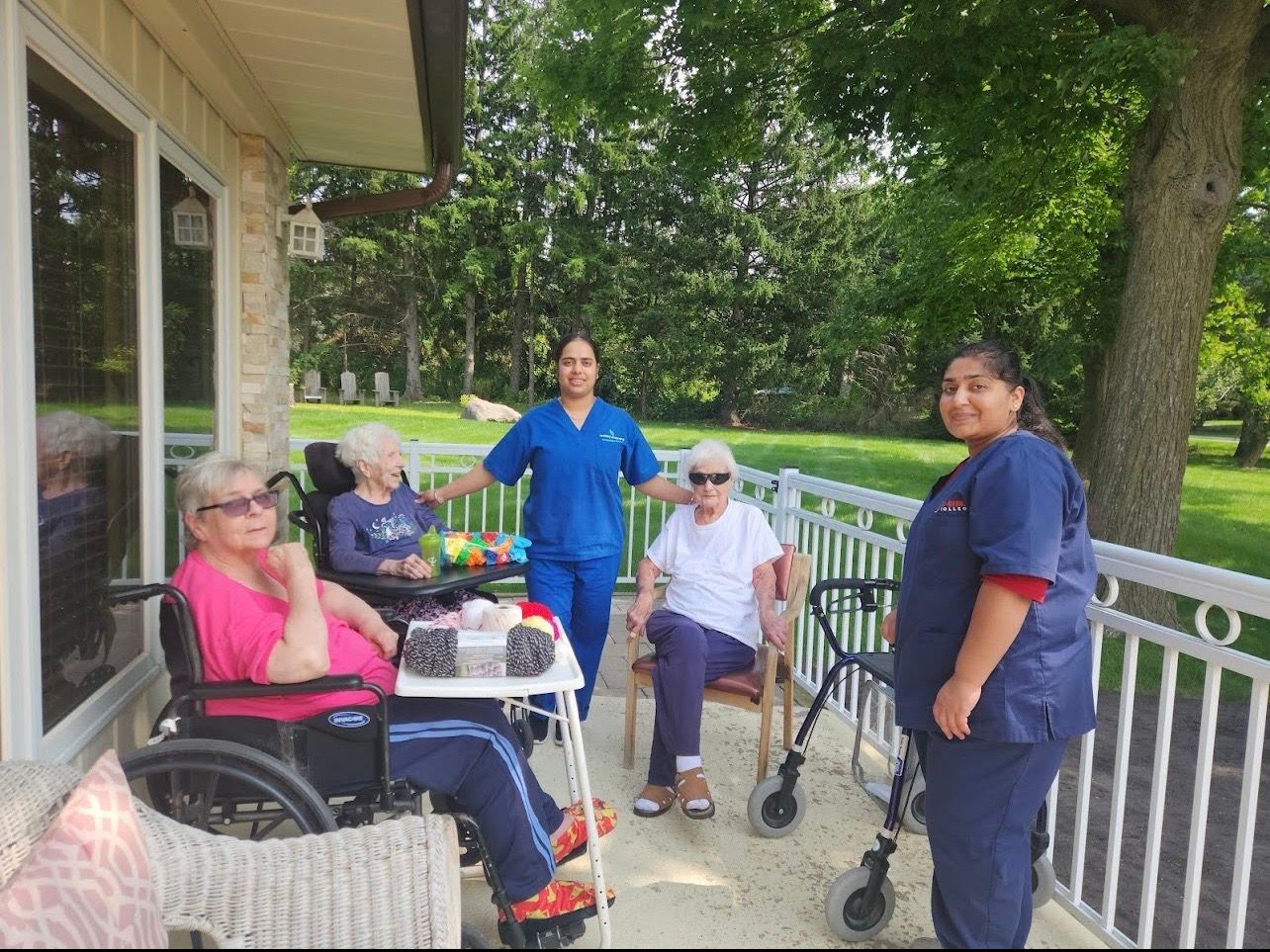 People seated on a porch with caregivers standing nearby, in a leafy outdoor setting.
