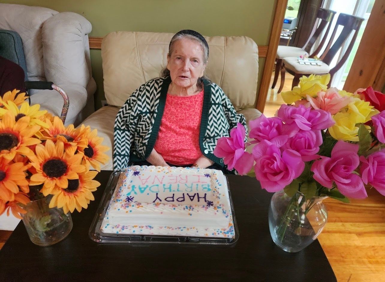 Woman seated at a table with flowers and a birthday cake, smiling in a living room