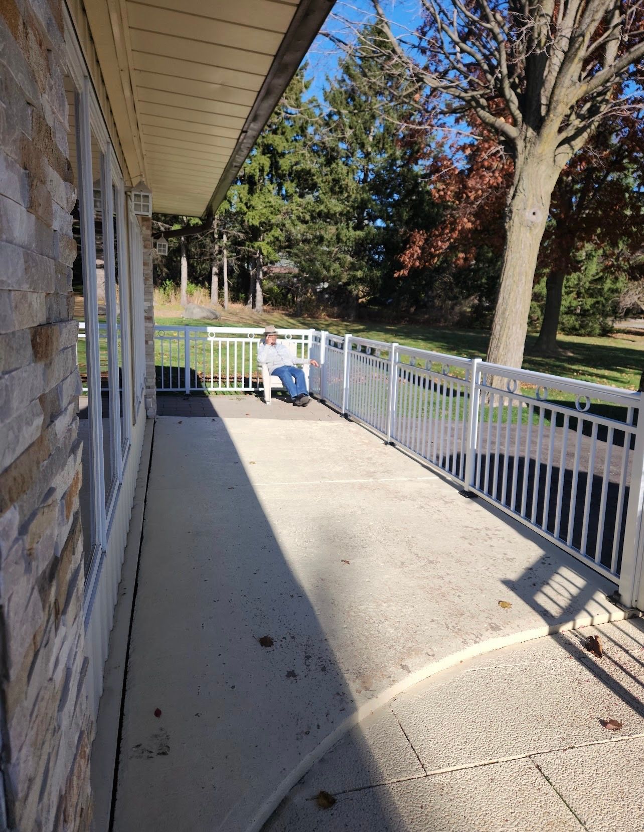 Sunlit porch with white railing beside a brick house, overlooking a yard and trees.