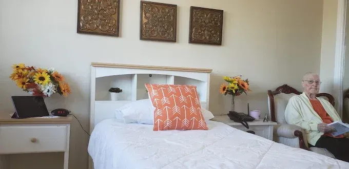 Hospital room with a white bed, flowers, wall art, and an elderly man seated in a chair by the window.