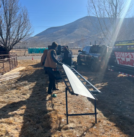 Two people installing a white panel outdoors near a truck and mountains on a sunny day.