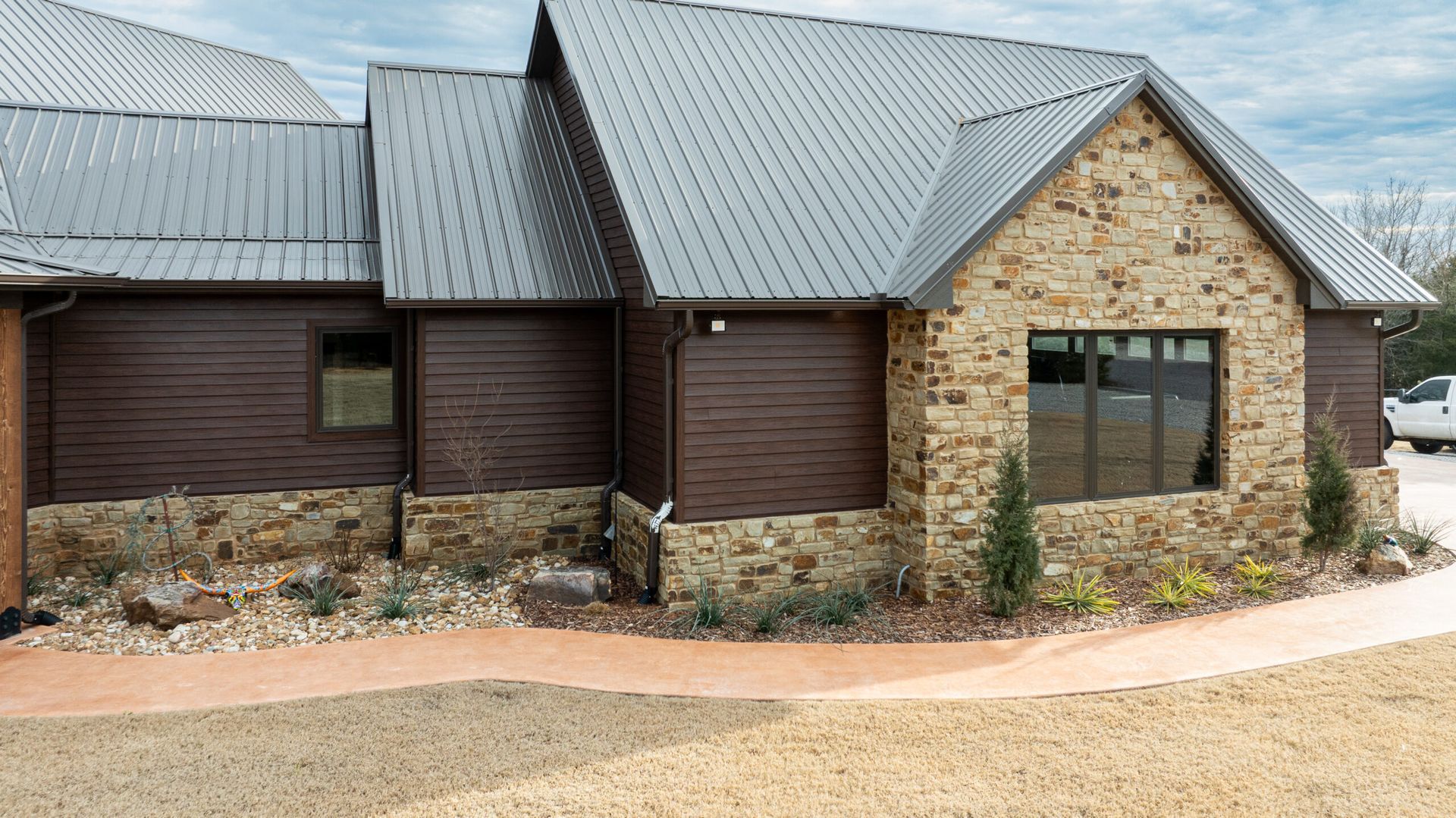 Stone-and-wood house exterior with metal roof and large windows, set along a gravel drive.