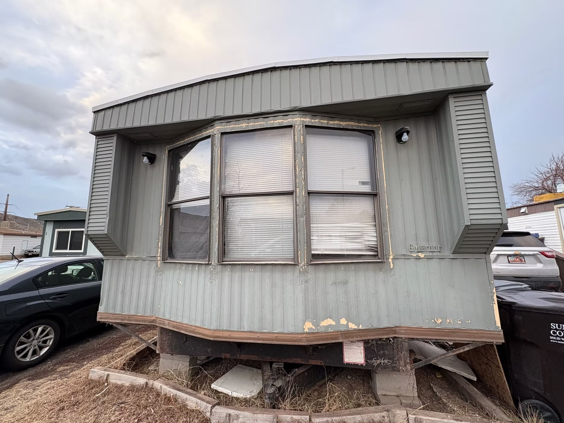 A weathered, gray mobile home with a bay window, set on blocks, with exposed undercarriage and surrounding debris.