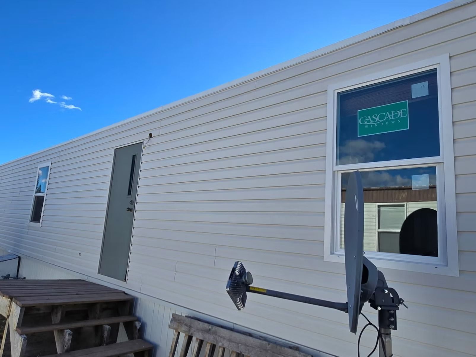 Exterior of a light-colored mobile home with a door, windows, and a satellite dish against a blue sky.
