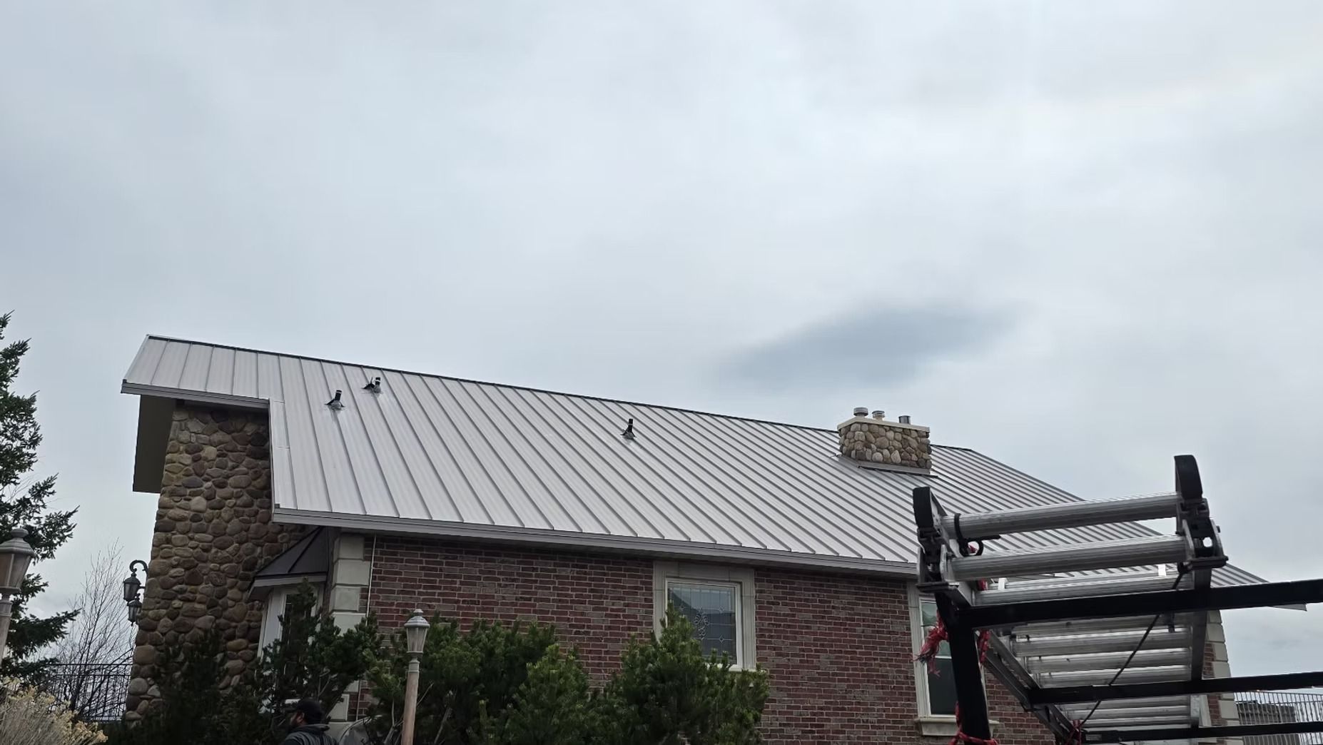 Brick house with a gray metal roof under an overcast sky, with wooden scaffolding on the right.