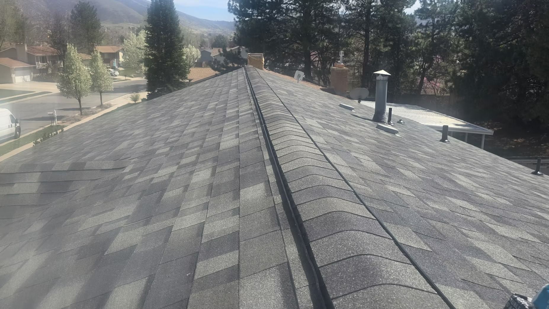 A roof with gray shingles, viewed from above. Ridge cap and chimneys visible, trees and houses in the background.