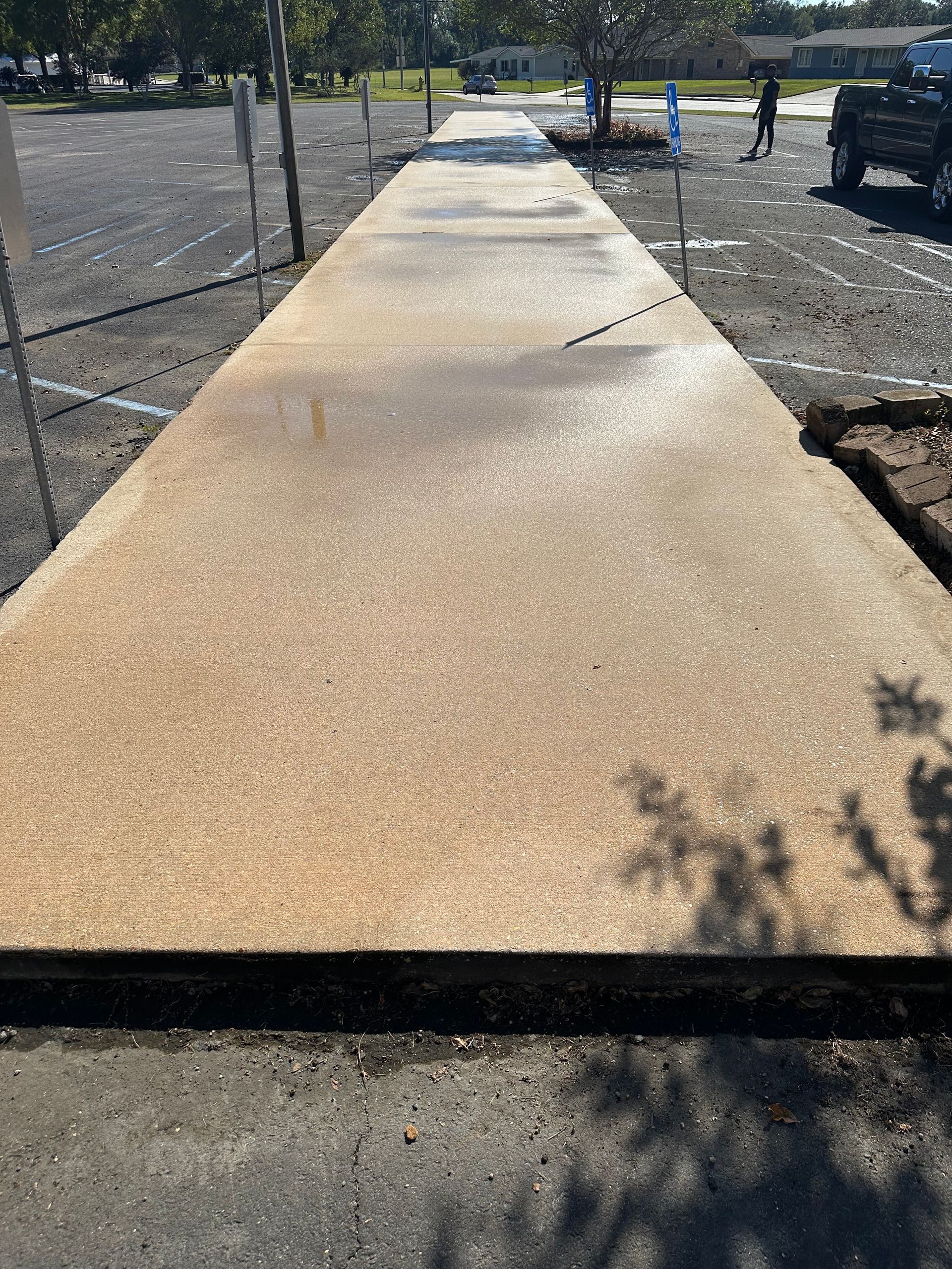 Concrete sidewalk, tan with aggregate, leading from parking lot, person walking in background.