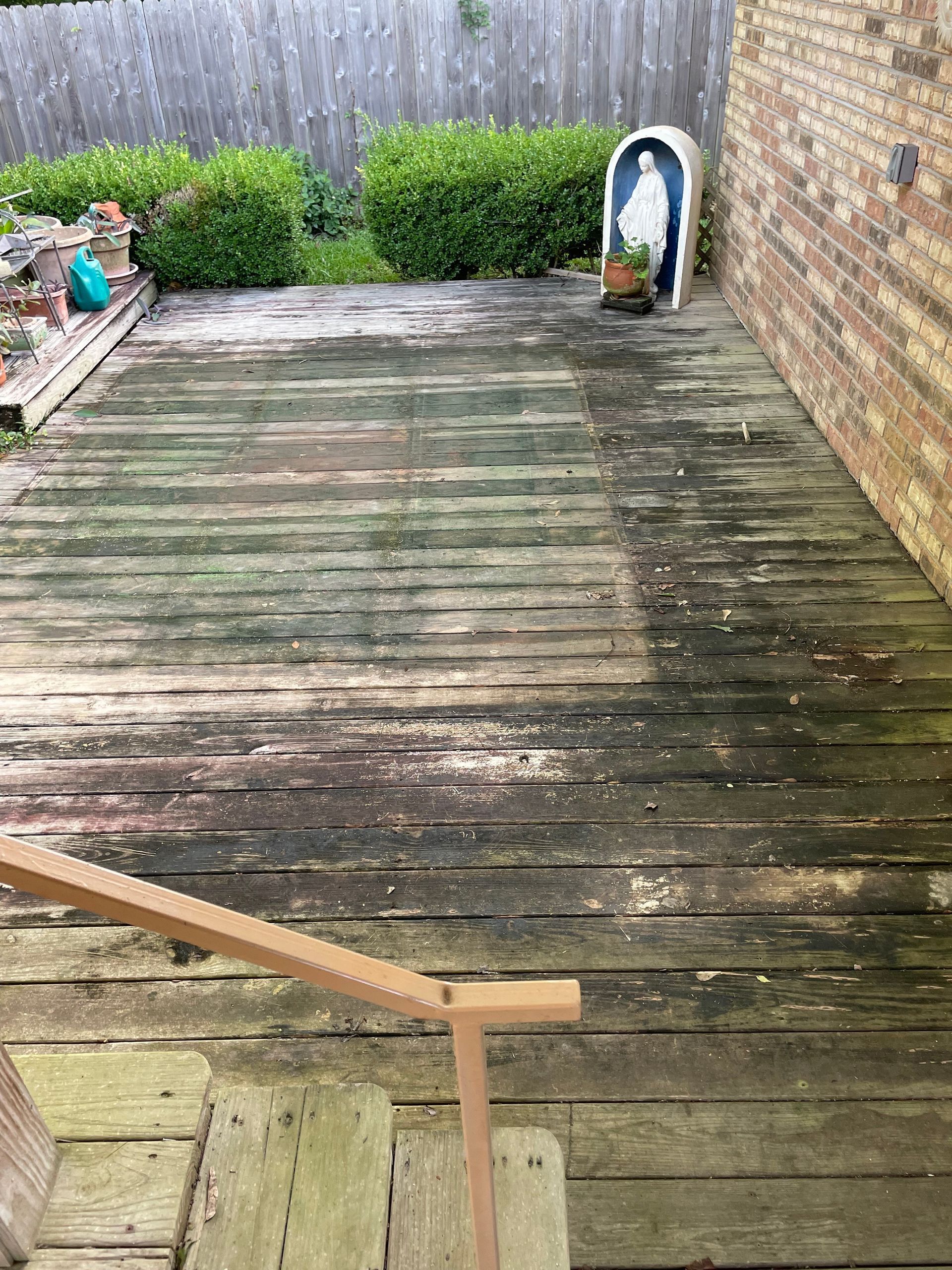 Worn wooden deck with a handrail, leading up to a yard, with a statue and bushes in the background.