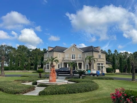 Large beige house with a fountain and manicured lawn under a blue sky with fluffy clouds.