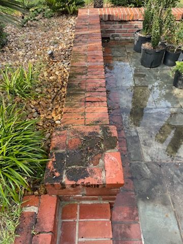 Brick wall with dark stains next to a paved area with plants and gravel.