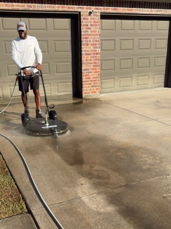 Person pressure washing a concrete driveway with a rotary surface cleaner near a garage.
