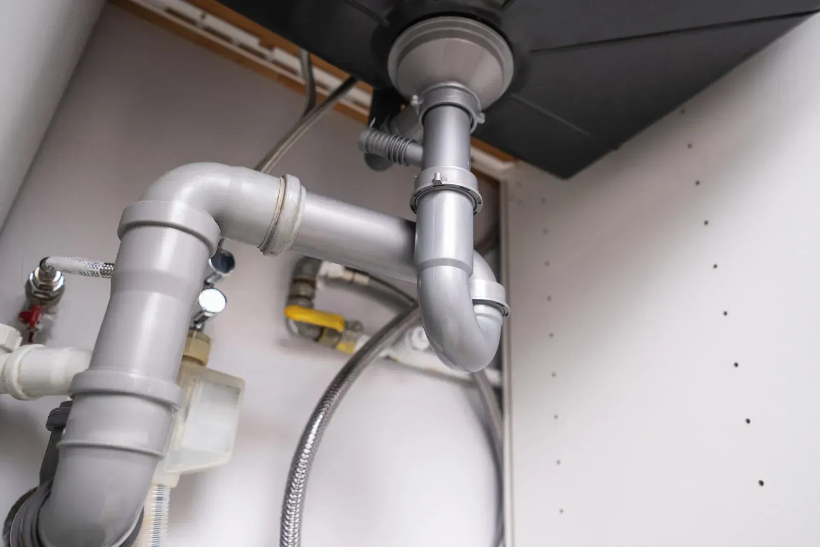 Grey plastic plumbing pipes, including a P-trap, connected beneath a kitchen sink inside a white cabinet.