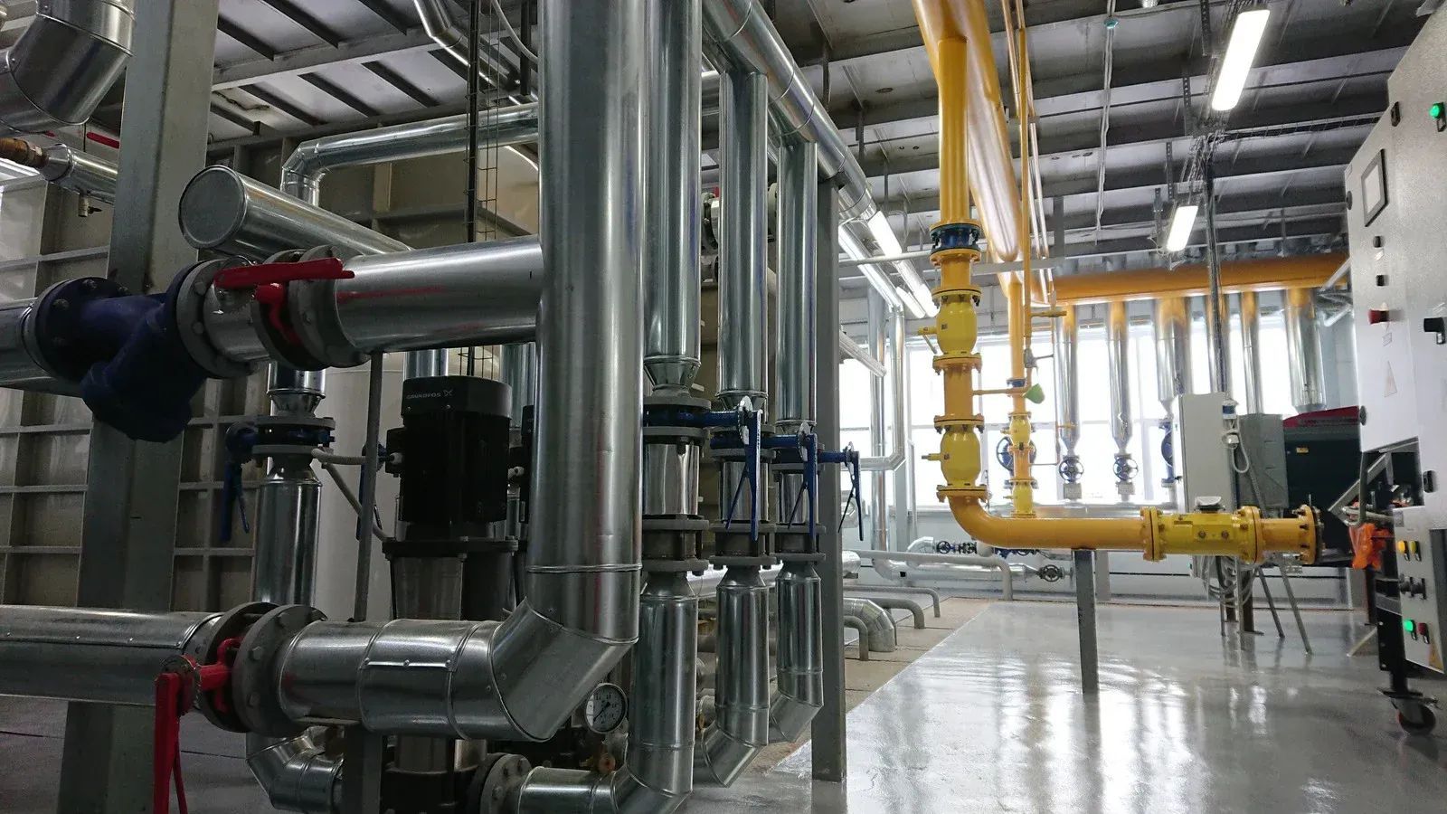 Industrial plant interior with large metal pipes, valves, and yellow overhead piping under bright lights.