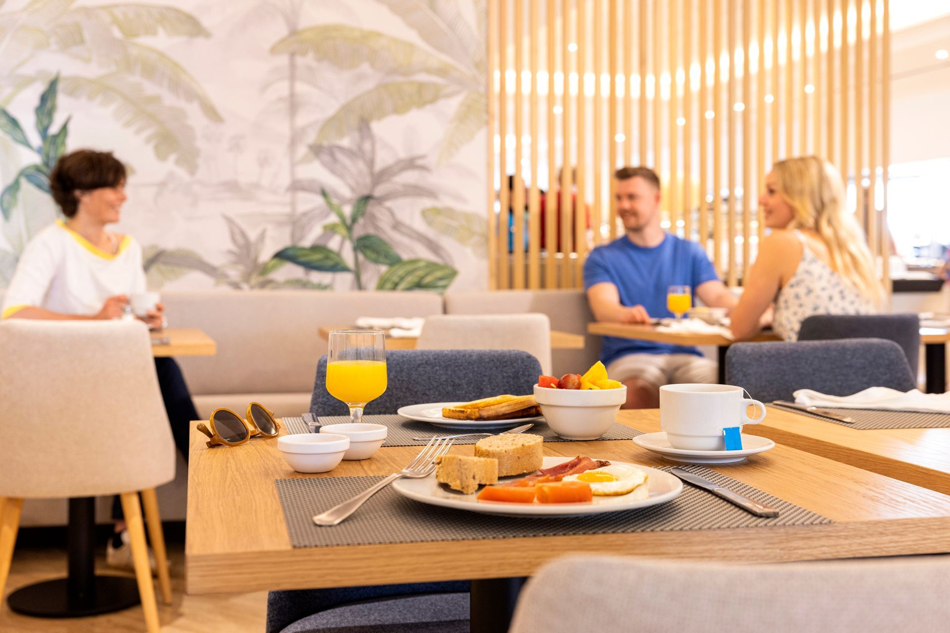People at a hotel breakfast. Table with food and drinks. Tropical mural in the background.