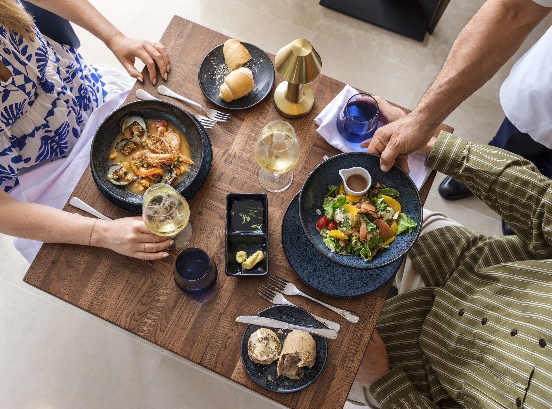 People dining at a table with various plates of food, drinks, and a small lamp.