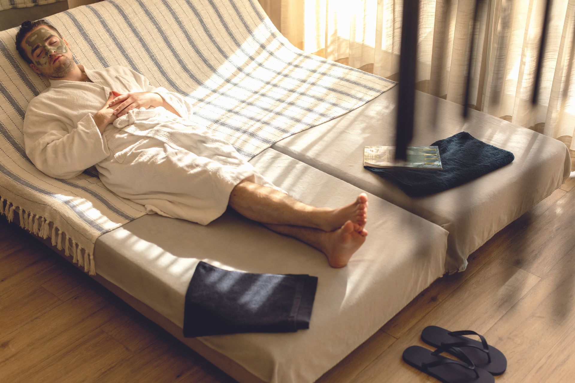 Man in robe relaxing on spa bed, face mask on, sunlit room.