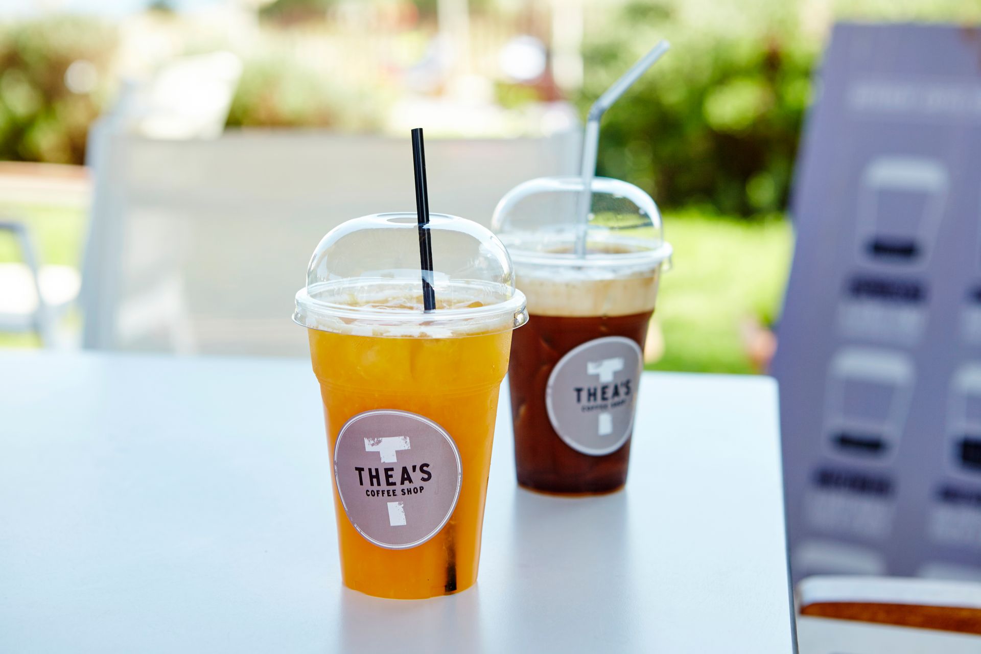 Two iced drinks with straws on a white table, outdoors. One orange, one brown.