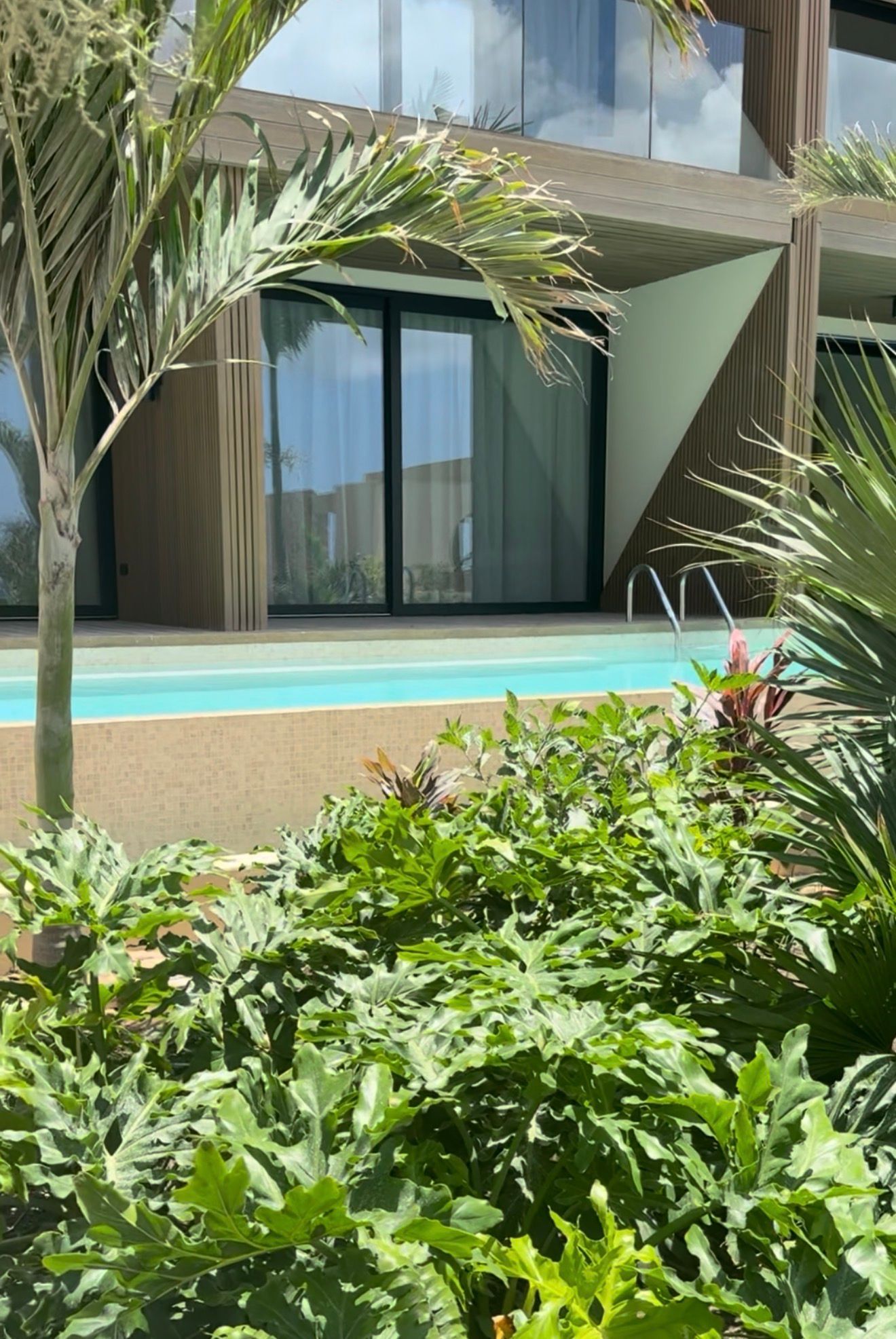 Hotelkamer met toegang tot het zwembad. Een houten terras met ligstoelen leidt naar een kamer met glazen schuifdeuren.