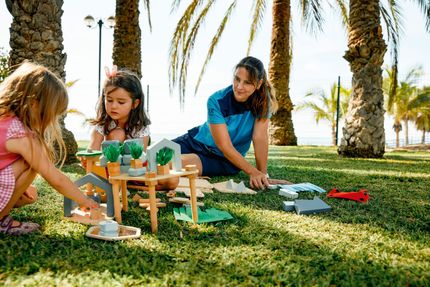 Woman and two girls playing with a wooden toy set in a park with palm trees.