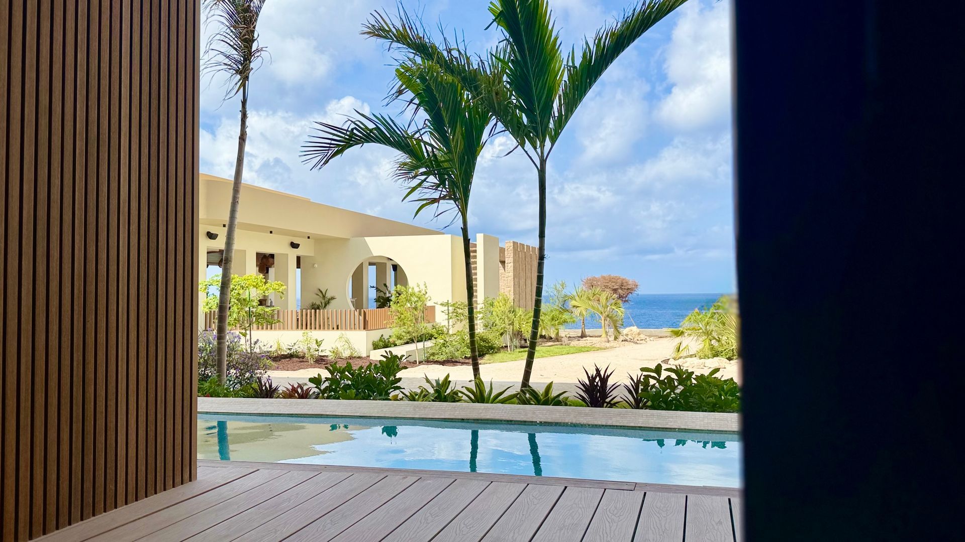 Lounge chairs beside a pool, facing hotel building with palm trees. Sunny day.